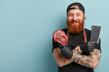 Image of satisfied red haired butcher smiles happily, involved in working process, keeps tattooed arms folded, carries cleaver and fresh meat, prepares pork for transporting, stands over blue wall