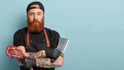 Studio shot of serious redhead manufacture worker, uses sharp cleaver for cutting raw meat, looks directly at camera, wears black protective gloves, cap and apron, isolated on blue background