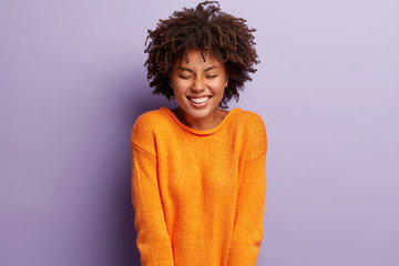 Candid shot of attractive Afro American woman laughs happily, keeps eyes shut from joy and pleasure, hears pleasant compliment, feels shy, dressed in orange outfit, stands indoor over lilac wall