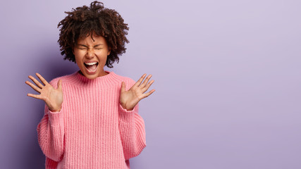 Image of emotional joyful Afro American woman spreads palms, has mouth wide opened, exclaims emotionaly, wears knitted jumper, glad to be promoted at work, has bushy black hair, poses indoor