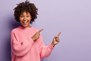 Waist up shot of positive lovely woman with Afro hairstyle, points away with both fore fingers, shows somethig on blank space, has joy expression, wears loose pink jumper, gestures over violet wall