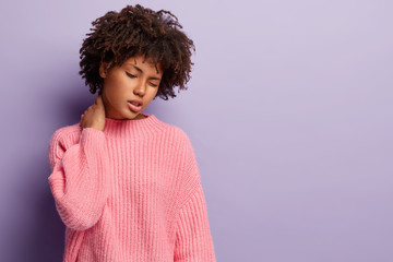 Displeased dark skinned lady has terrible pain in neck, suffers from osteochondrosis, has desk work, sad expression, wears oversized pink jumper, isolated over purple background. Tiredness concept