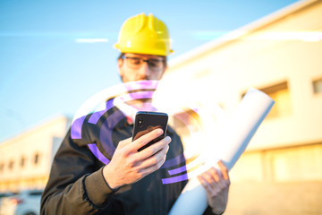 Giovane ingegnere con giacca nera, occhiali da vista e caschetto giallo sta interagendo con il suo smartphone digitale e connessione wifi per dare ordini alla squadra di operai.