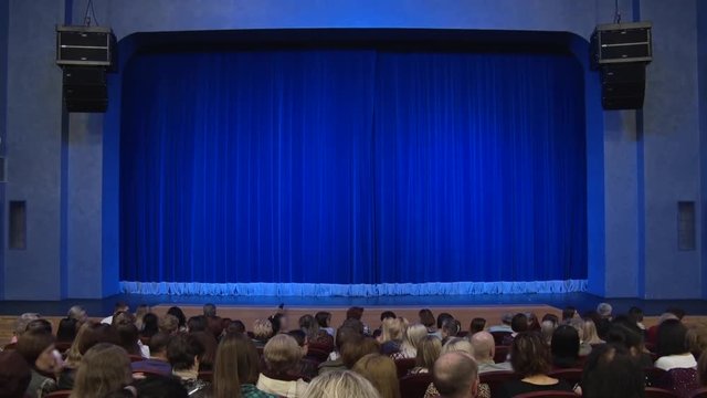 People in the auditorium of the theater before the performance or in the intermission. Blue curtain on stage. Shooting from behind.