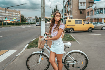 Fototapeta na wymiar The girl summer city at intersection, stands with a bicycle, waiting for the traffic light to turn on the road. Pink blouse white shorts, on the handlebars bag shopping bag from the store.