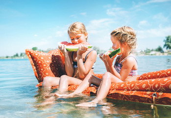 Two little sisters sitting on innflatable mattrace and eating watermelon