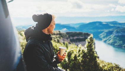 hipster tourist hold in hands mug of hot drink, lonely smile guy enjoy sun flare mountain in auto,...