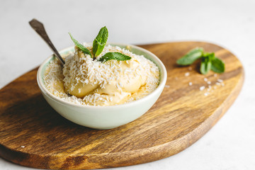 Homemade vegan ice cream in a bowl is made from frozen mango and coconut milk served by fresh mint and flakes on a wooden textured board.