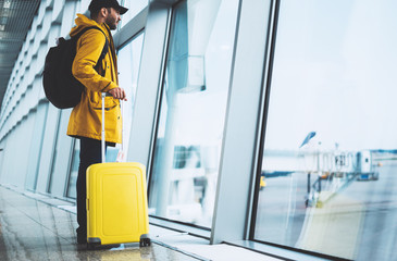 traveler with yellow suitcase backpack at airport on background window blue sky, passenger waiting flight in departure hall of lobby terminal lounge area, vacation trip concept, empty space mockup
