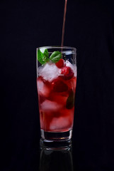 Red drink with ice, raspberries and mint in a tall glass against the black background
