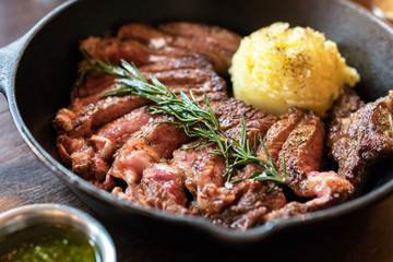 Cast iron plate with sliced grilled rib-eye cooking a medium-rare steak with mashed potato and sauce in saucer over table.