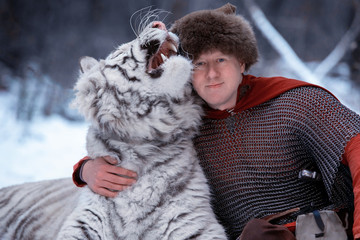 Man dressed as a medieval warrior sits in the snow in a winter forest and hugs a white tiger by the neck. The warrior is dressed in chain mail, a fur hat and a red cloak. - Powered by Adobe