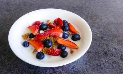 Vibrant closeup of white breakfast bowl with cereal of flakes, coconut & banana, topped with blueberries and strawberries, glistening with fresh cow's milk. Dish on a black marble in natural light.