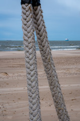 two braided heavy ropes in close-up; In the background there is a sea sandy beach and a sea with a large ship