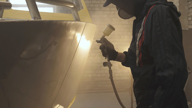 In slow motion a professional man (guy) in a garage (service) in working clothes dressed in a sweater sets up a paint gun (spray gun) and starts painting (putting a layer of ceramics) a yacht.