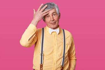 Close up portrait of mature man posing with hand on forehead isolated over pink background. Handsome elder man posing in photo studio, dressed yellow tshirt in one tone, suspenders and white bow tie.