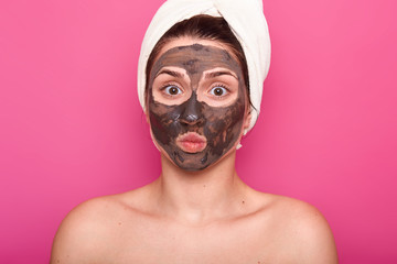 Close up shot of beautiful female, has chocolate mask on face, keeps eyes wide opened and lips rounded, poses with naked body, has white towel on head, having rest in spa salon, isolated on pink wall.