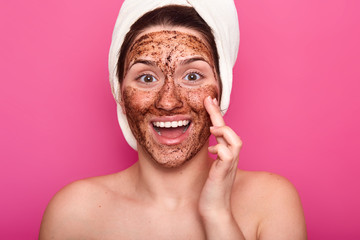 Isolated shot of surprised woman applies mask on face, has happy facial expresion, opens mouth from amezmant, having rest while doing beauty procedures, wears white towel on head. Wellness concept