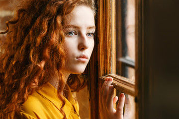 Pensive curly ginger girl in the yellow shirt looking to the window at the loft interior
