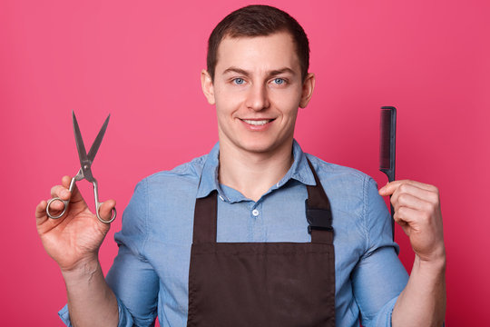 Positive handsome brunett barber shows scissors and comb, dressed in blue shirt and brown apron, ready to do hairstyle for men, poses smoling against pink background. Grooming concept. Copy space.