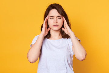 Image of young good looking brunette female stands with closed eyes isolated over yellow background, keeps hands on temples, has awful headache. Charming girl wears white t shirt. Healthy concept.