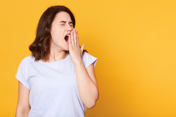 Portrait of tired sleepy girl in white casual t shirt yawning isolated on yellow background, stands...
