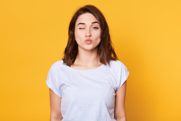 Close up portrait of adorable brunette woman pouts lips and winks, makes grimace, rounds lips for...