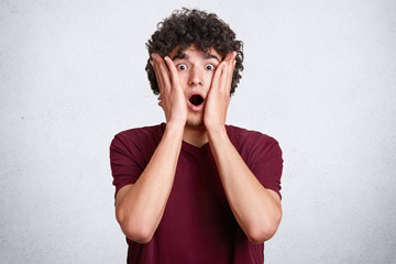 Horizontal portrait of handsome Caucasian young guy with astonished facial expression, wears casual maroon t shirt, touches cheeks, keeps open mouth, hears shocked news, isolated on white background.