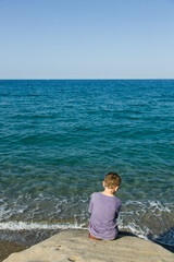 Fototapeta na wymiar Blue sky and green - blue sea, horizon, silent waves and a little sitting boy can be seen from behind, in foreground coastal rock.
