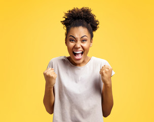 Excited girl with widely open mouth holds hands clenched in fists, exclaimed with positiveness. Photo of african american girl on yellow background. Emotions and pleasant feelings concept.