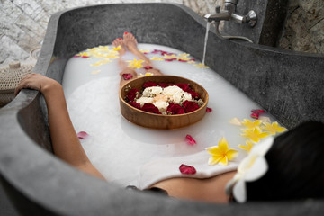 Young woman relaxing in black stone bath with tropical flowers and rose petals. Skin treatment, luxury spa concept.