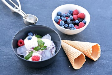 A dark bowl full of wild berry ice cream balls with mint leaves and ice cream waffles next to it on a dark kitchen surface - concept with refreshing fruit ice for the summer