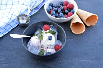 A dark bowl full of wild berry ice cream balls with mint leaves and ice cream waffles next to it on a dark kitchen surface - concept with refreshing fruit ice for the summer