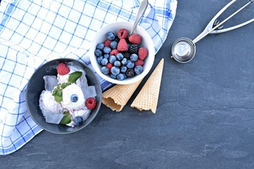 A dark bowl full of wild berry ice cream balls with mint leaves and ice cream waffles next to it on a dark kitchen surface - concept with refreshing fruit ice for the summer