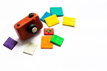A small vintage camera of brown color is made of plasticine. Next to the camera is a few photos of bright and colorful flowers. The whole composition lies on a white background.
