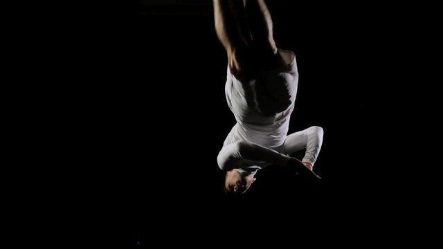 Athletic man in white clothes performs tricks jumping on a trampoline on a black monophonic background