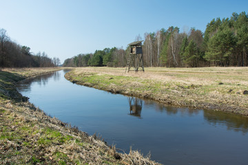 Obraz premium Quiet river flowing through a meadow, a hunting pulpit in the forest and blue sky
