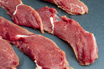 Appetizing red fresh veal, sliced into raw thin beef steaks, laid out on a black stone slate. The...