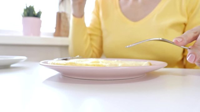 Woman sitting by the table, eating an omlet with a folk and a knife and drinking coffee or tea for breakfast at home or restaurant