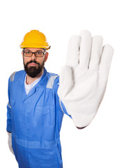 Fun high angle of bearded workman with yellow helmet, blue uniform and in glasses making stop gesture with his hand in gloves. over white background