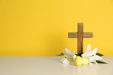 Wooden cross, Easter eggs and blossom lilies on table against color background, space for text