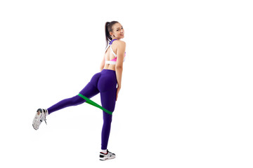 A ypung woman coach in a sporty purple  short top and gym leggings makes lunges  by the feet forward with sport fitness rubber bands,  stretch legs on a  white isolated background in studio