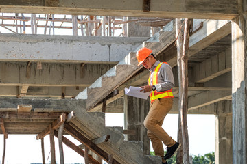 Asian architect or engineer holding looking project blueprint papers and wear helmet at construction site. He walking on stairs.