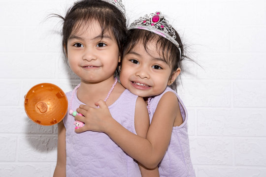 Female Asian Identical Twins Sitting On Chair With White Background. Wearing Purple Dress And Accessories. Hugging Each Other While Standing