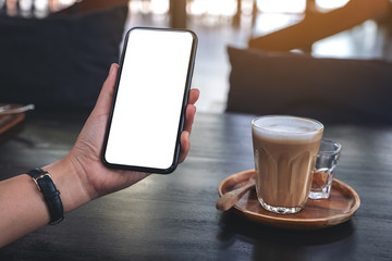 Mockup image of a woman's hand holding black mobile phone with blank screen with a glass of coffee on wooden table in cafe