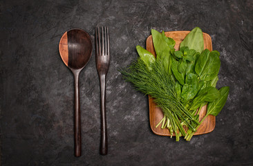 Naklejka na ściany i meble Fresh herbs – spinach, sorrel, dill on wooden plate with wooden salad fork and spoon on black background . Vegan, diet and clean eating concept.