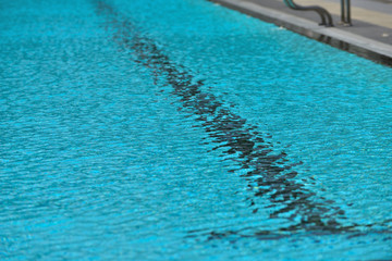 A swimming pool with a sunny reflection and beautiful wavy water surface in turquoise blue color with grab bars ladder on the sides and black line lanes underwater.