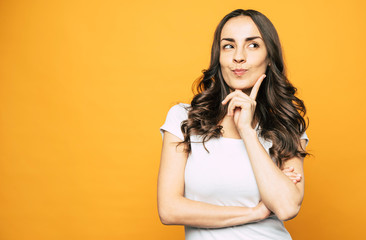 Let me think. Musing nice-looking girl next to the orange background in basic outfit is thinking about something and we can see it through her gestures and mimic.