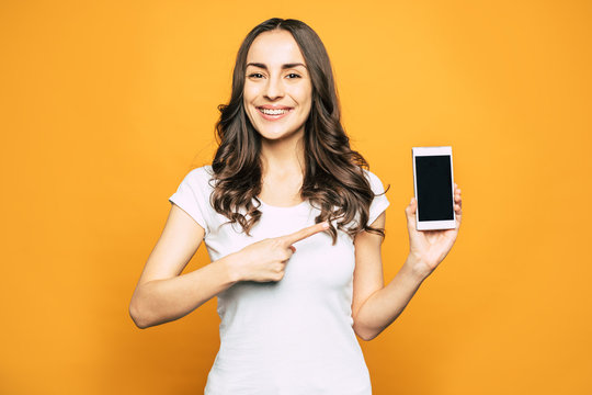 Note That. Glad Pretty Girl Is Pointing Right On The Phone In Her Hand With Riant Smile On Her Beautiful Face In Front Of Spice Orange Background.