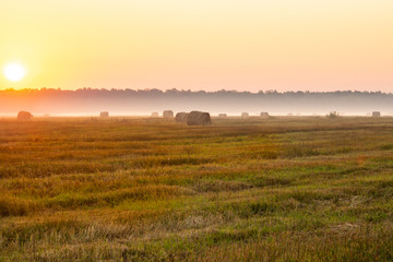 Rising sun dispersing morning mist in the field with bales of hay
